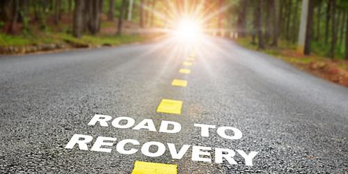 The words 'road to recovery' printed on the middle of a road, with sunshine in distance